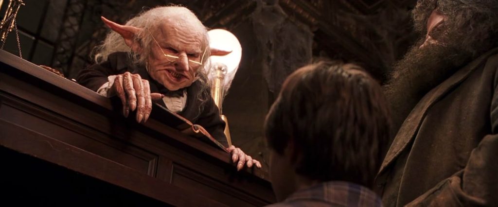 A goblin wearing gold half-rimmed glasses and an old-fashioned black suit sneers over a counter brown wood at Harry Potter and Hagrid.