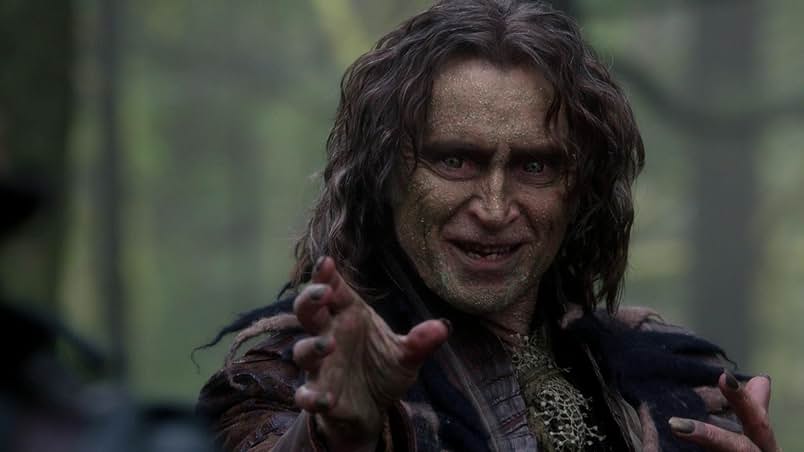 A man with wavy hair and a gold, scaly face sneers as he reaches forward. Behind him is the blurred background of a forest.