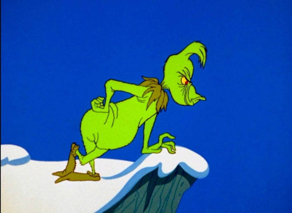 A dark blue sky with a snowy cliff. Learning over a snowdrift is the sneering Grinch. His fist is on his hip, and he taps his finger on the snow.
