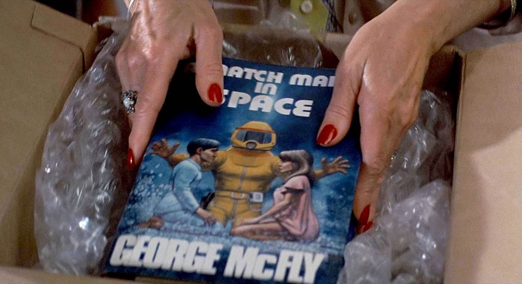 Hands with red nail polish and a big diamond ring reach into a cardboard box full of bubblewrap. Within the bubblewrap is a copy of the book A Match Made in Space. The book cover has a blue background with a figure in a yellow hazmat suit reaching out towards a boy and girl in pajamas facing each other on their knees.