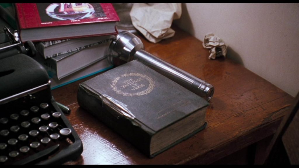 A cluttered school desk with balled up paper, a stack of four books, a silver flashlight, and a black typewriter. Beside the flashlight is a worn black book with a taped spine. Five Centuries of Verse is in golden lettering with a golden wreath around it.