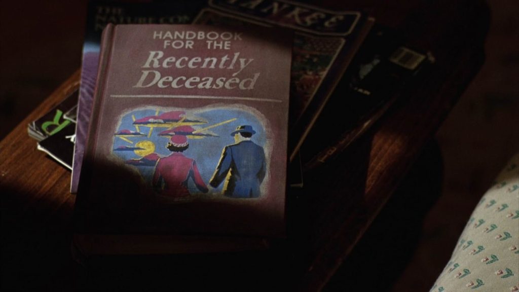 A wooden side table full of magazines is cast in shadow. Resting on top of the magazines is a copy of the book Handbook for the Recently Deceased. It has a brown background with a man in a blue suit and hat and a woman in a pink suit and hat holding hands and facing the sky. There is a bright yellow sun and clouds tinged with pink and blue.