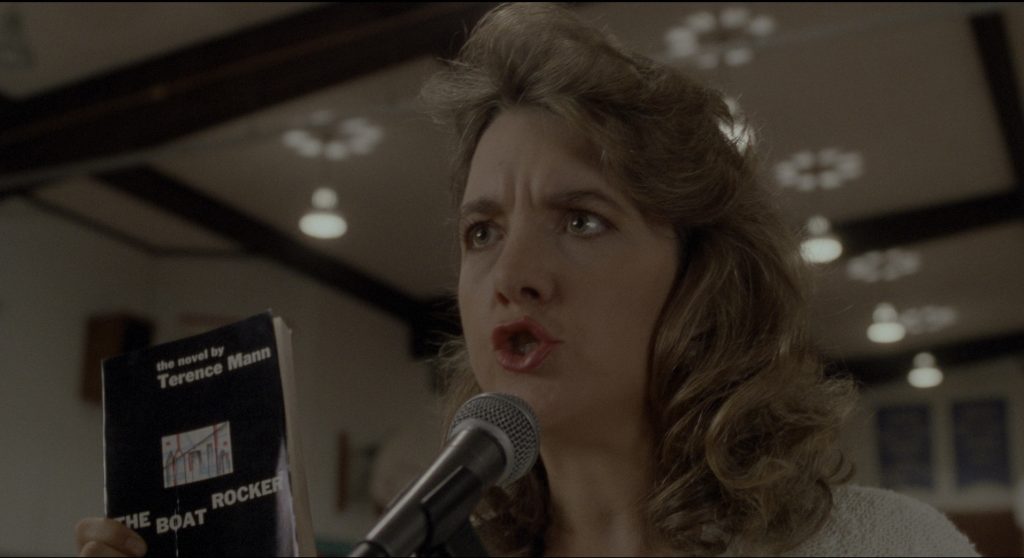 A woman speaks angrily into a microphone and holds a copy of the book The Boat Rocker. The book has a black cover with a small white, red, and blue sketch of a city skyline.