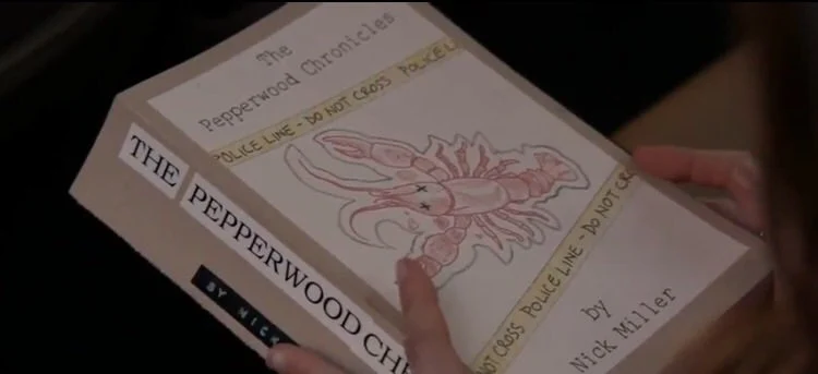 Hands hold a tan copy of the book The Pepperwood Chronicles. On the front cover is a white background with a red sketch of a dead lobster and yellow crime scene tape.