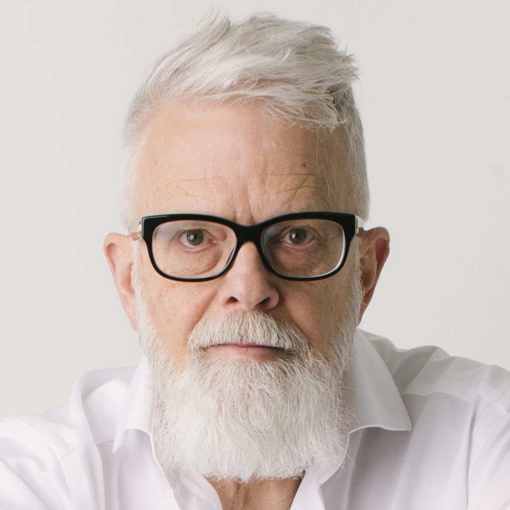 David Hayward, a light-skinned man with short, straight white hair and a white beard and mustache, wearing black-framed glasses and a white shirt, posing in front of a white background.