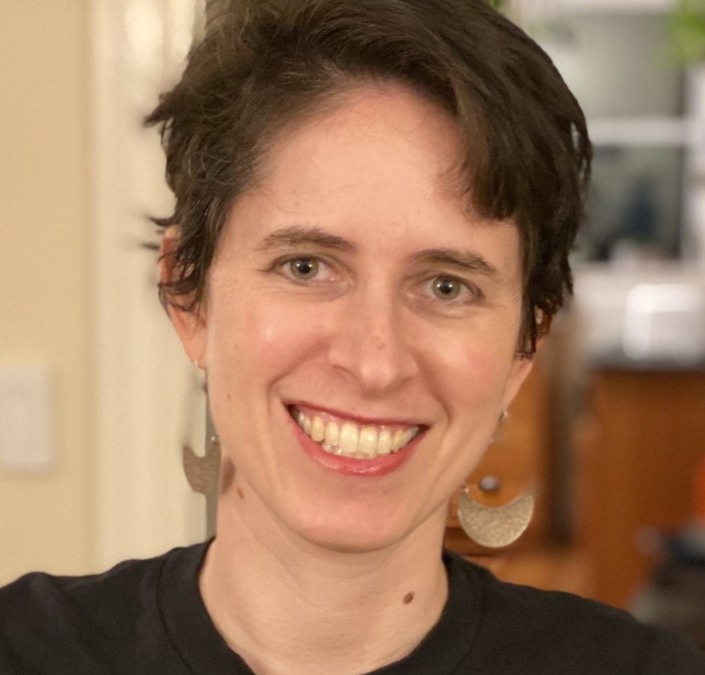 Rabbi Shoshana Meira Friedman, a smiling light-skinned woman with short brown hair and green eyes, wearing a black t-shirt and dangling half-moon-shaped earrings.