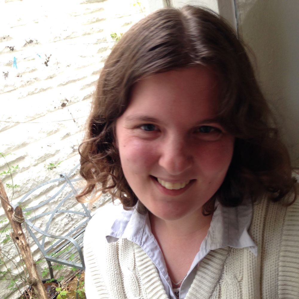 Rebecca Burgess, a smiling light-skinned person with shoulder-length curly brown hair and blue eyes, wearing a blue collared shirt under a white sweater vest, sitting next to a window that overlooks a garden.