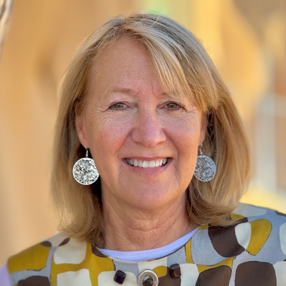 Jeanne Walker Harvey, a smiling light-skinned woman with shoulder-length, straight blonde hair and hazel eyes, wearing round silver earrings and a brown, yellow, and gray spotted blouse, standing in an outdoor area that is largely warm-toned.