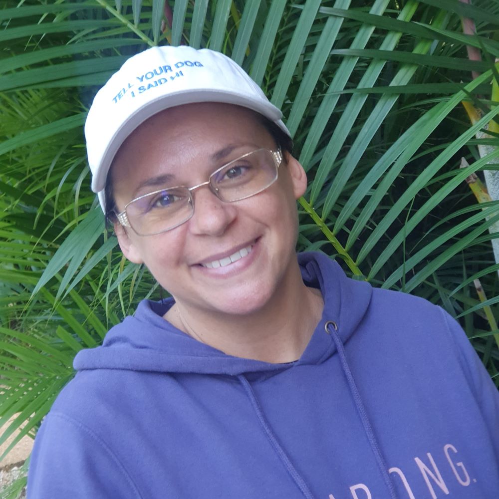 Kate Foster, a smiling tan-skinned woman with hazel eyes, wearing a white ball cap that says "Tell Your Dog I Said Hi" and a blue hooded sweatshirt, standing in front of a wall of leafy palm plants.