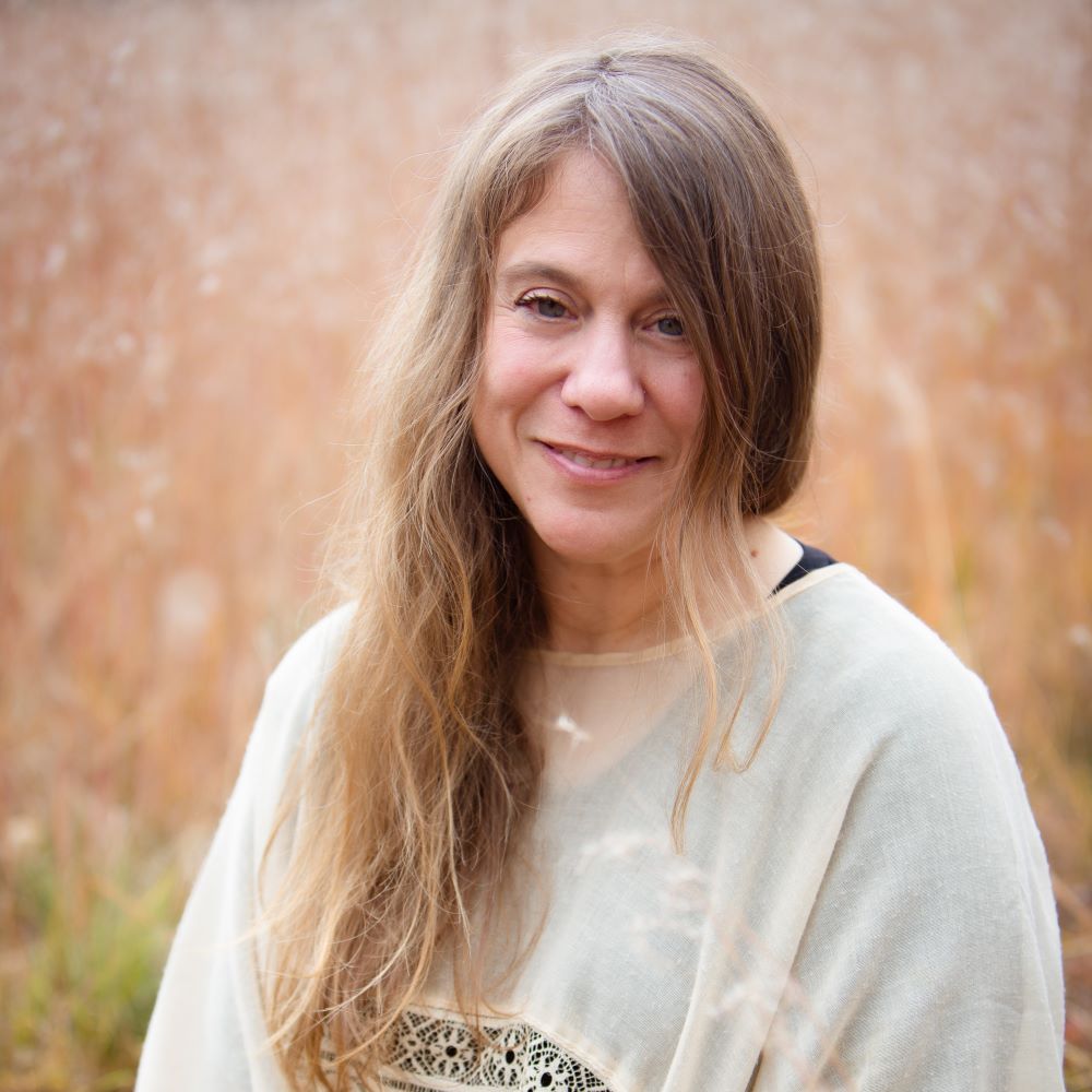 Marcella Pixley, a light-skinned woman with long dark blonde hair and blue eyes, wearing a white blouse while posing outside in a field of orange grasses.