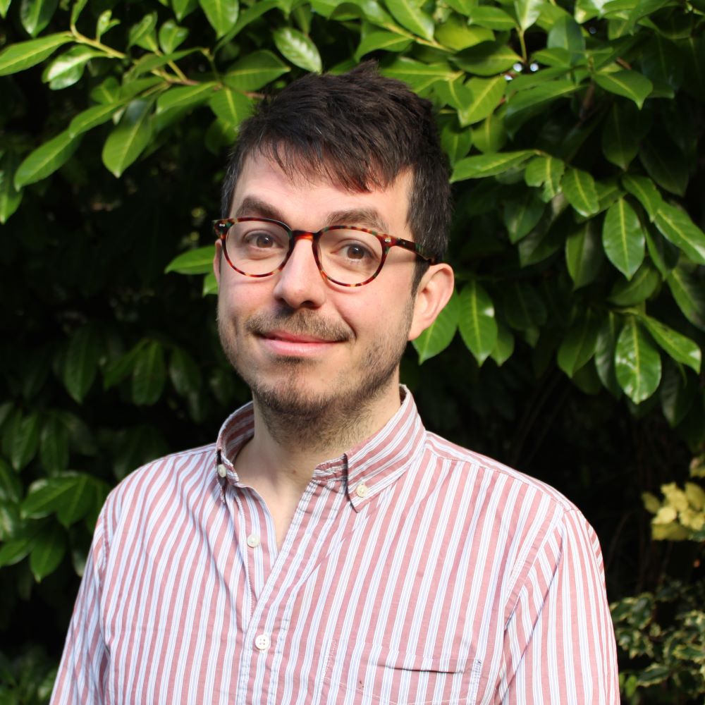 Ross Montgomery, a light-skinned man with short, straight dark hair and brown eyes, wearing tortoiseshell glasses and a pink and white striped button-down shirt, standing in front of a tall leafy plant.