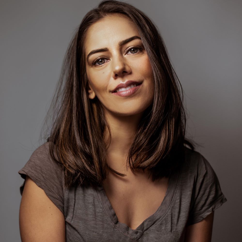 Sheetal Sheth, a smiling brown-skinned woman with shoulder-length, straight dark brown hair and hazel eyes wearing a gray t-shirt against a gray background.