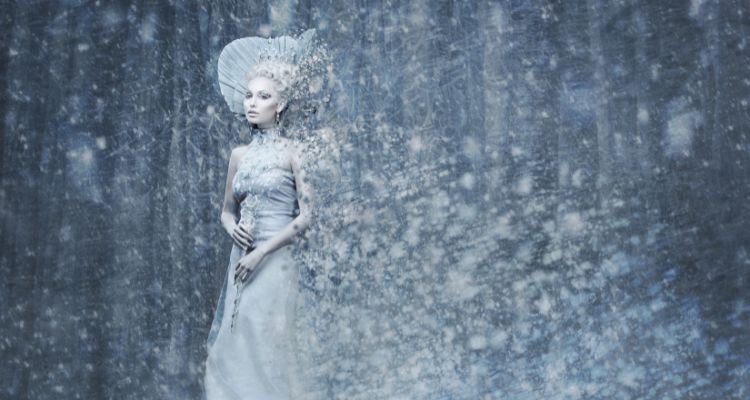 A white-skinned adult with white-blond hair and wearing an elegant white gown stands in the center of a snowstorm, with bits of ice and snow seeming to emerge from her body.