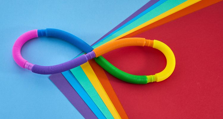 A rainbow set of accordion mobility toys fashioned into the shape of an infinity symbol rests on a pile of multicolored papers and a blue surface.
