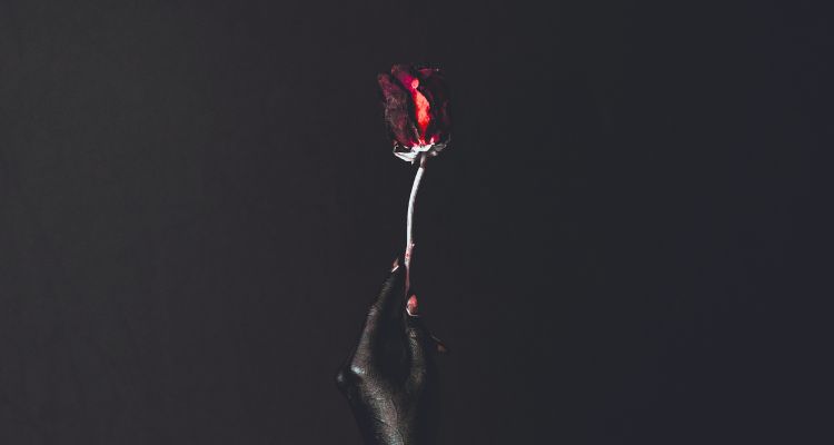 A hand with unnaturally black skin and red-clawed fingertips holds a red rose that is beginning to wilt against a dark background.