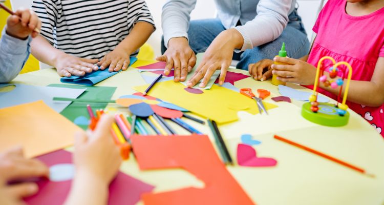The hands of an adult and a group of small children work together on paper crafts at a round table; art supplies like glue and colored pencils are on the table as well.