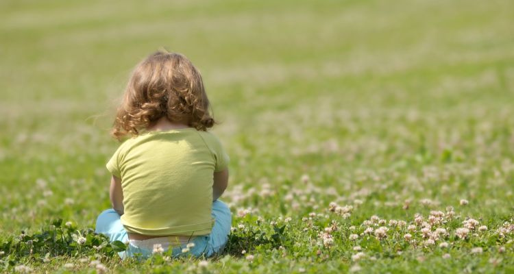 A light-skinned child with shoulder-length, wavy brown hair sits in a grassy field with small white flowers with their back facing the viewer.