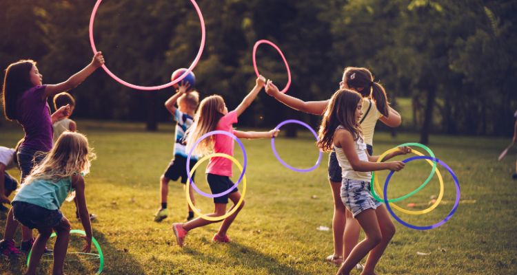 A group of children diverse in skin tone and hair color play with hula hoops, throwing rings, and balls outside at sunset.