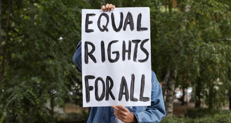 A brown-skinned adult holds a white sign with black writing that reads "Equal Rights for All," obscuring their facial features, in an outdoor, tree-filled area.