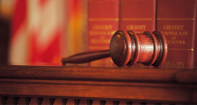 A wooden gavel rests on a wooden surface; a row of law books' spines are visible behind it.