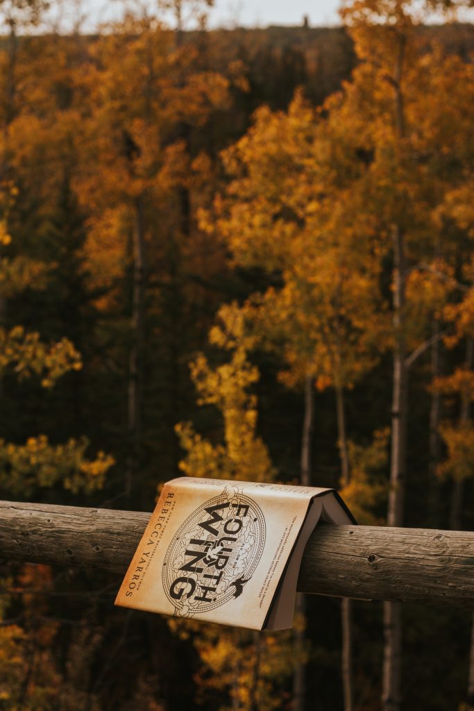 A section of a wooden pole fence with a copy of the book Fourth Wing resting on top. Behind is a forest full of trees with orange and yellow leaves.