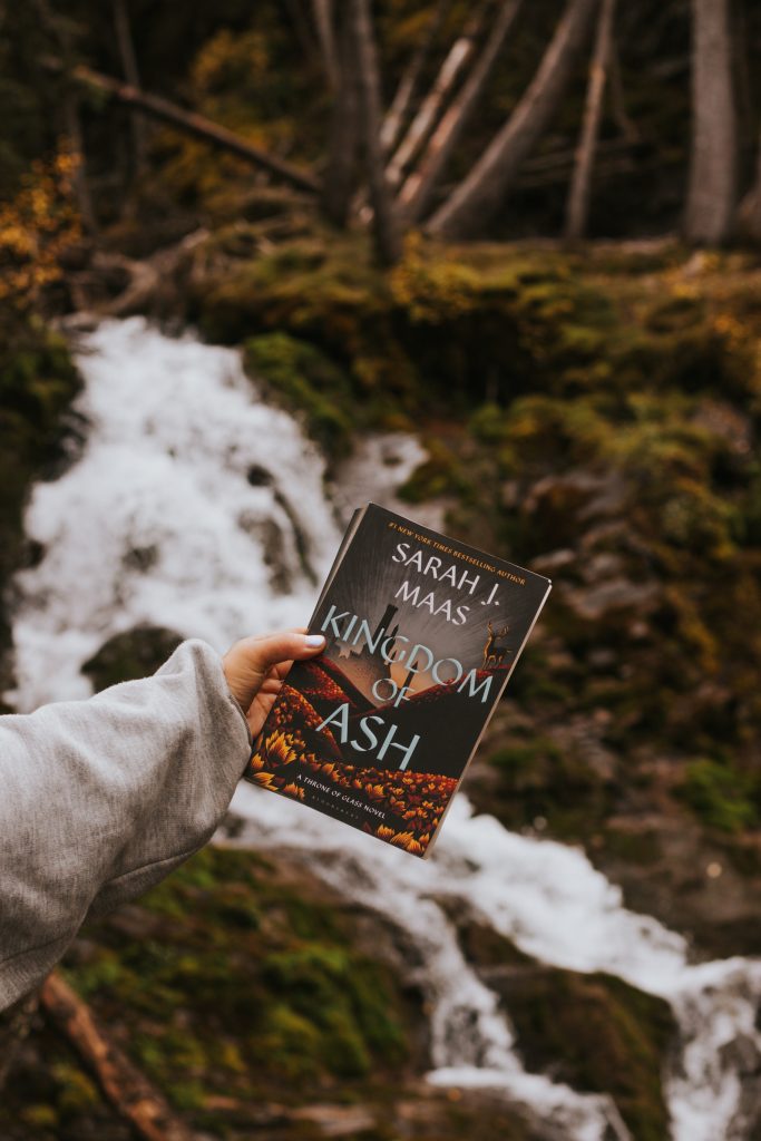 A hand wearing a gray sweatshirt holds a copy of the book Kingdom of Ash. Behind, is a forest hill with mossy terrain and a small waterfall.