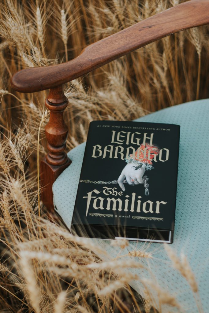 A chair with dark wooden arms and a blue cushion seat. Resting on the chair is a copy of the book The Familiar. Behind is a wheat field.