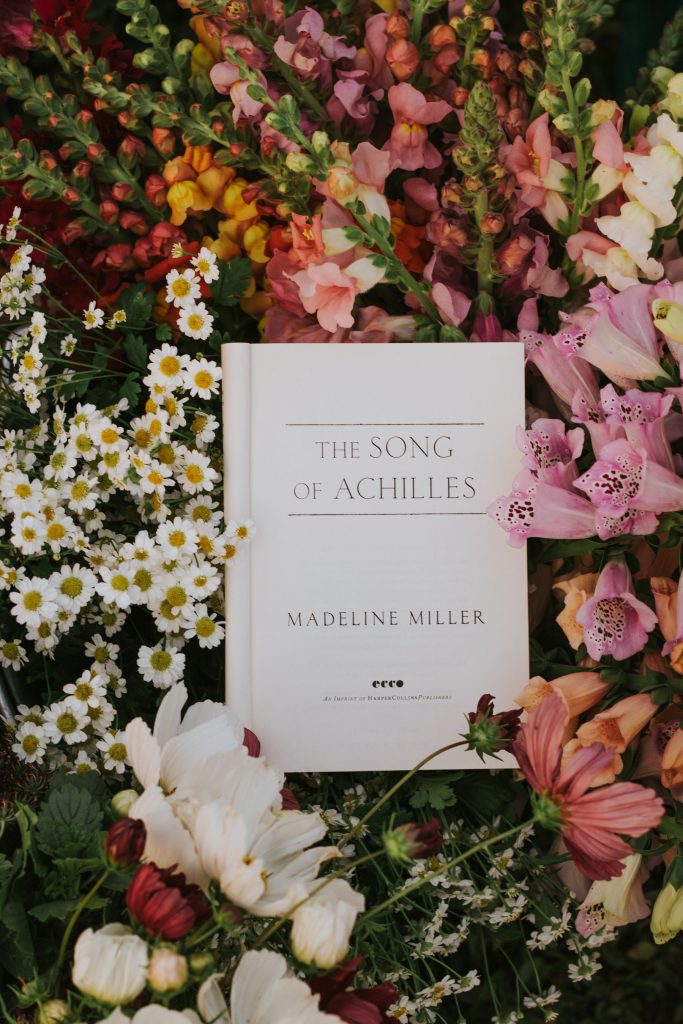 A copy of the book The Song of Achilles resting in a pile of white, pink, red, and yellow flowers.