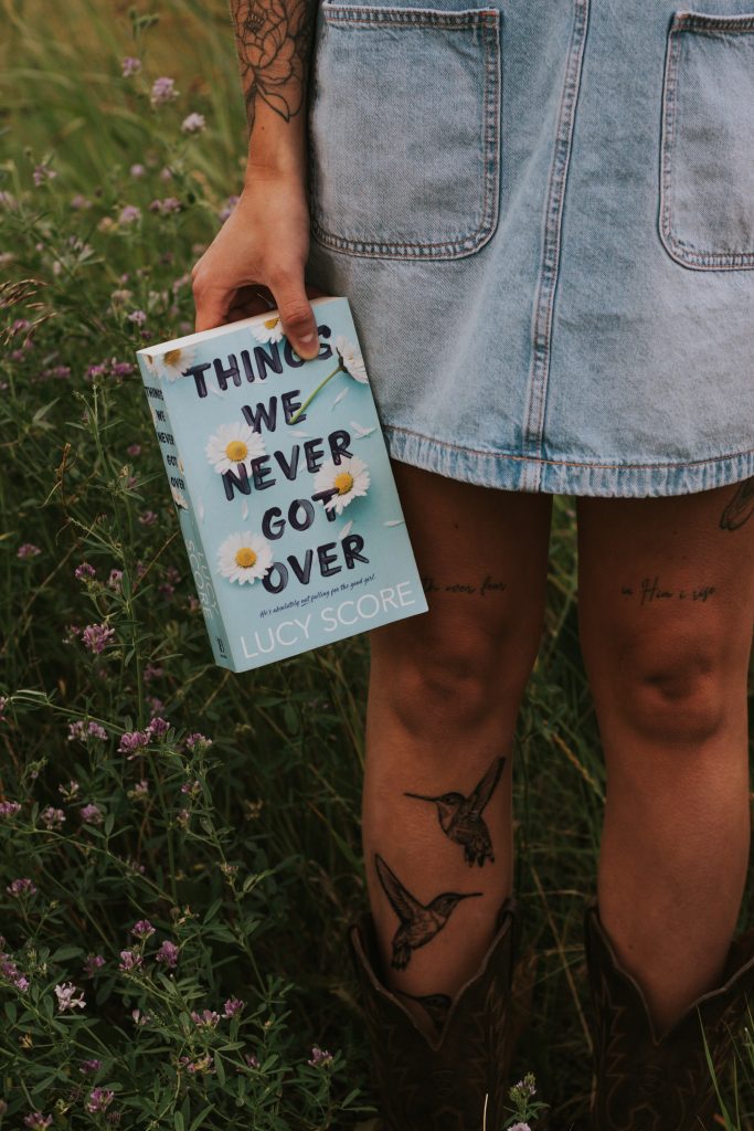 A woman's tattooed legs. She wears a light blue denim skirt and stands in a field of purple wildflowers. In her hand is a copy of the book Things We Never Got Over.
