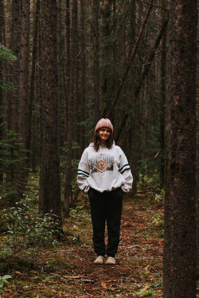 A woman in an orange, pink, and blue striped beanie, a white Yellowstone hoodie, and black pants stands in a forest.