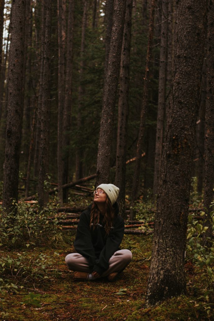 A woman in a white beanie, glasses, a black hoodie, and brown pants sits cross-legged in a forest and looks to the left.