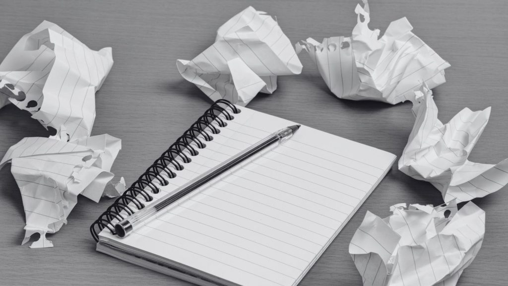 Grayscale photo of a spiral-bound lined notebook with a pen on top, lying on a desk, surrounded by crumpled notebook pages.