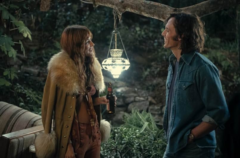 A woman has a camel fur-lined jacket draped over her shoulders and wears a burgundy tied midriff with matching bottoms. She smiles and holds a beer bottle. Across from her is a smiling man wearing a denim shirt and a blue t-shirt. Behind them are a glowing Moroccan lamp, a brown striped couch, and stone steps leading into the woods.
