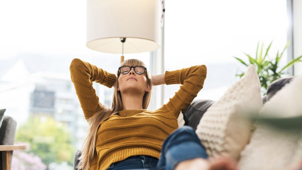 A light=skinned woman with long blonde hair reclines across a couch with her eyes closed and her hands behind her head. She wears glasses, a caramel-colored sweater, and jeans. Beside her are gray and white pillows. Behind her is a white lamp, a plant, and a large window with a view of buildings and a tree.