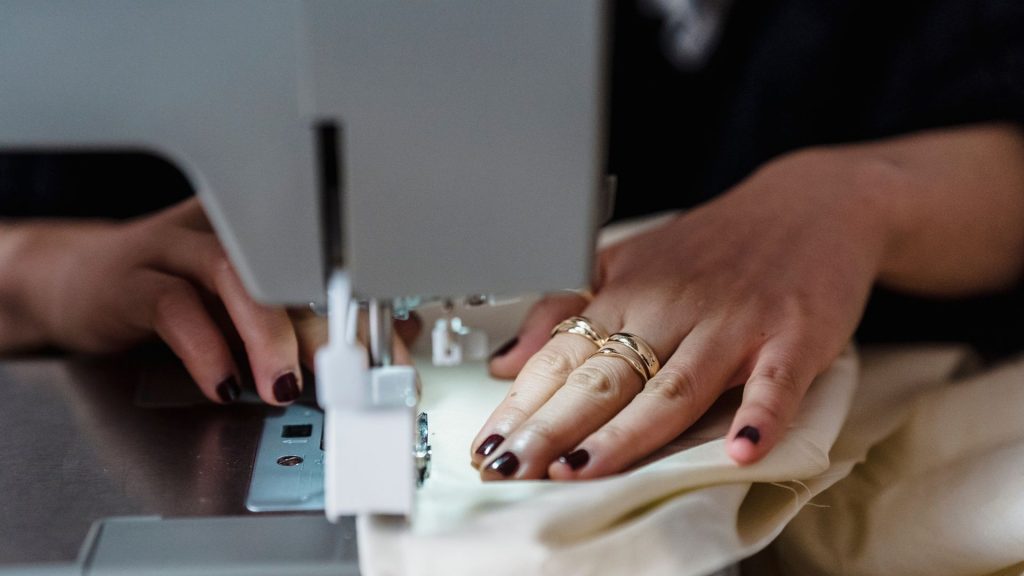 Two hands with dark nail polish sewing a white piece of fabric with a white sewing machine. The left hand has three gold rings, one on the index finger and two on the middle finger.