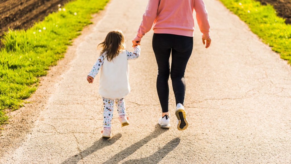 A mother and child walk down a cracked pathway lined on either side with wildflowers, grass, and dirt. The child has shoulder length brown hair and wears a dress with a long-sleeved shirt, leggings, and pink boots. The mom's head and shoulders are out of frame. She wears a pink sweater, black leggings, and white sneakers.