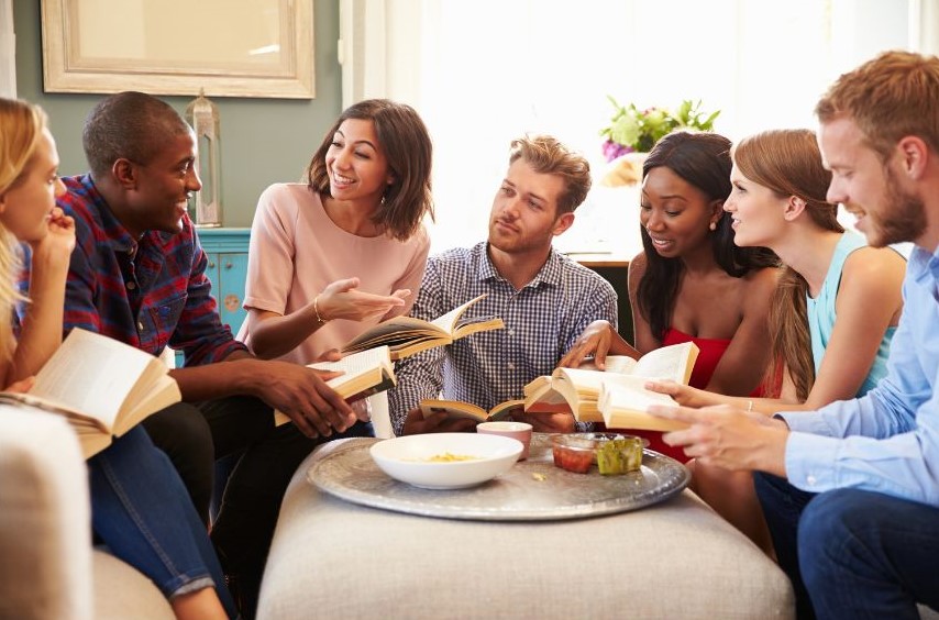 A group of seven people of genders, various skin colors, hair textures, and clothes sitting around a table that has a few snacks. They're all holding books and seem to be having an animated conversation.