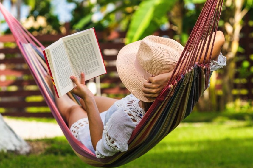 Someone lying on a hammock outside. wearng a gardening hat, holding up a book.