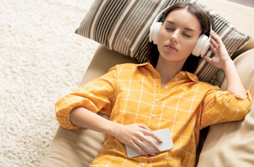 A woman lying on a light brown sofa with her head on a striped pillow, and her eyes are closed and headphones on, holding her phone on her stomach.