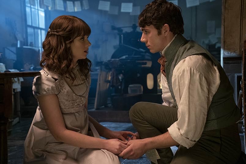 A man and a woman hold hands and crouch on the floor of a printer's shop filled with blue lighting. The woman wears a purple Regency era dress and the man wears a white shirt, gray vest, brown trousers, and a red cravat.