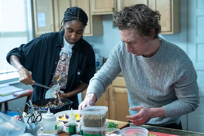 Two people cooking in a small kitchen with white walls and pale wood cabinets. On the left is a Black woman wearing a white graphic t-shirt and black button down shirt. On the right is a white man wearing a gray cable knit sweater.