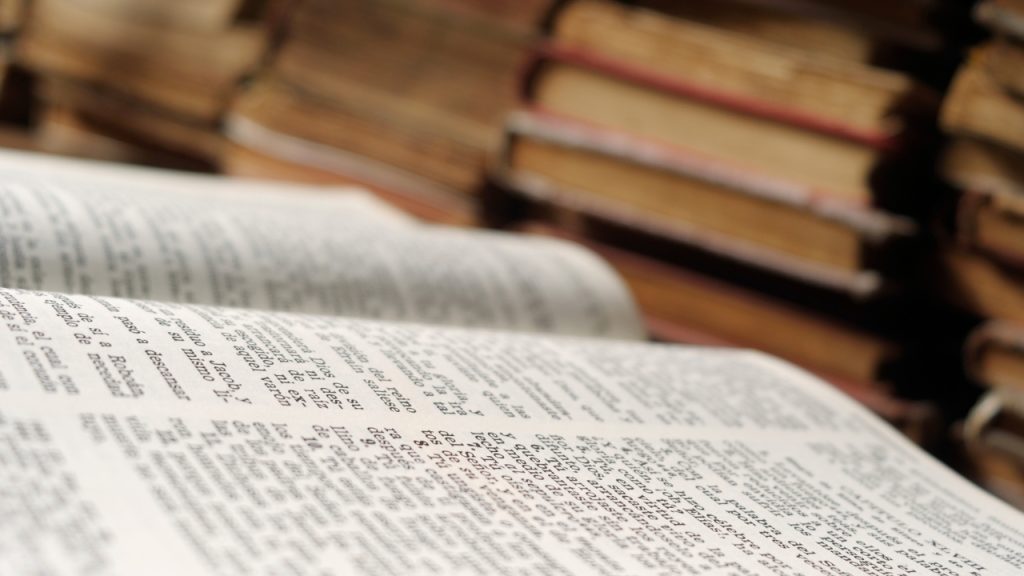 A close-up of an open book. In the background are blurred stacks of books.