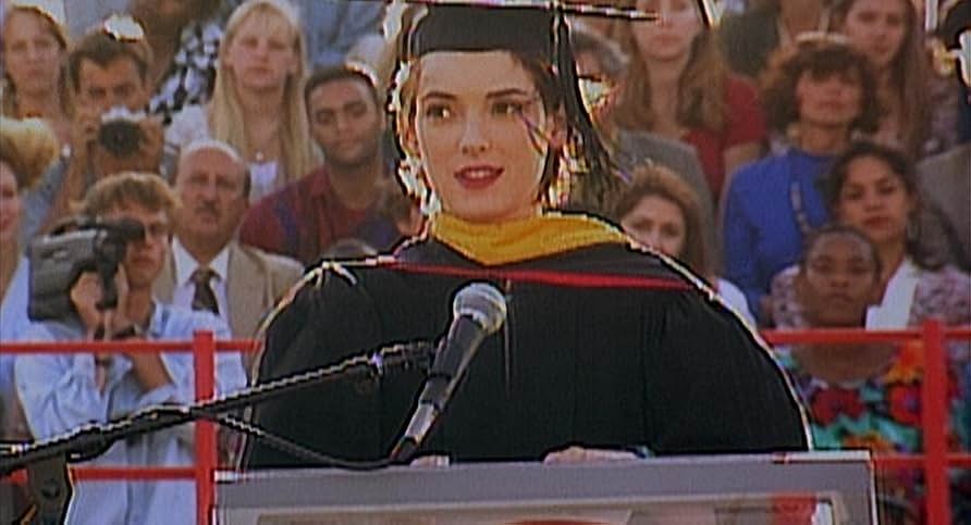A young woman in a black graduation cap and gown and yellow valedictorian stole stands at a podium with a microphone. Behind her is an audience of people behind a red railing.