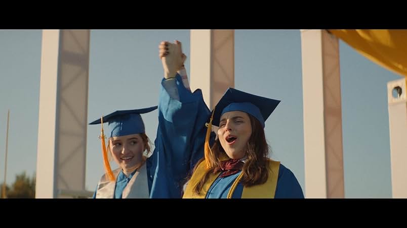 Two young women wear blue caps and gowns with yellow tassels. The woman on the left wears a white stole, and the woman on the right wears a yellow stole. They raise their clasped hands in the air and smile in celebration. Behind them are four makeshift white columns and a yellow canopy.