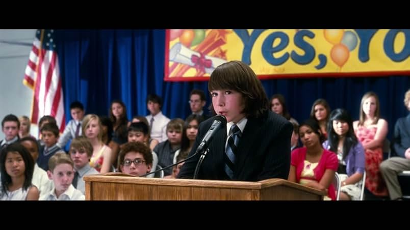 A boy wears a black suit, white shirt, and blue and black striped tie. He stands at a wooden podium and speaks into a microphone. Behind him is a seated audience of other teens in front of an American flag, blue curtains, and a yellow congratulatory banner.