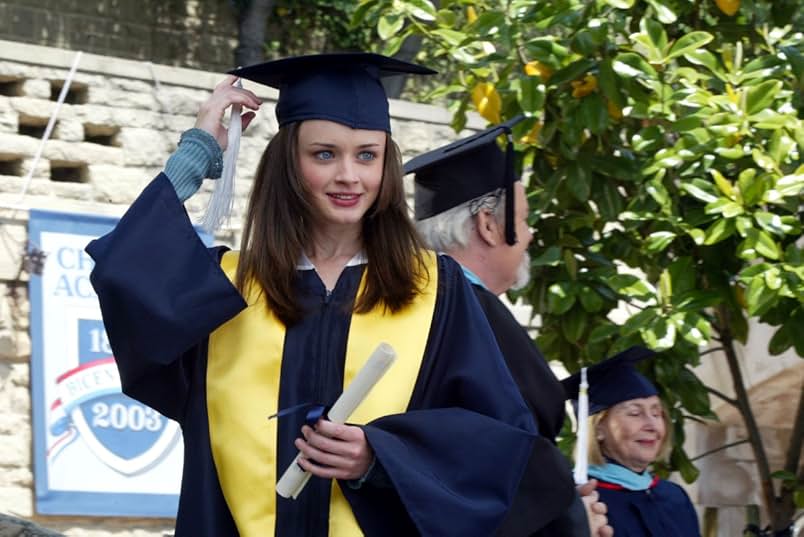 A young woman wearing a blue graduation cap and gown with a white tassel and yellow valedictorian stole holds a rolled diploma in her hand and reaches for her tassel. Behind her is a stone wall with a Chilton Academy banner, two teachers, and a tree.