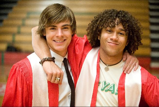 Two young men stand with their arms around each other's shoulders. They both wear red graduation gowns and white stoles. The man on the left wears a white button shirt and black tie under his gown. The Black man on the right wears a white t-shirt under his gown and a black cord necklace, brown bracelets, and a class ring. Behind them are gym bleachers.