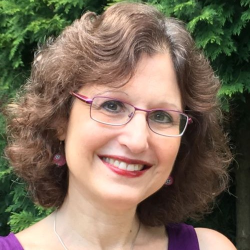 Jacqueline Jules, a light-skinned woman with short, wavy brown hair, smiles at the viewer. She wears pink rectangular glasses and stands in front of a tree.