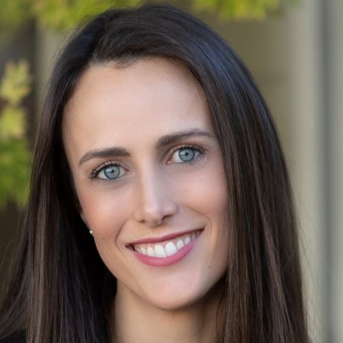 Charlotte Offsay, a light-skinned woman with long, straight brown hair, smiles at the camera. She stands in front of an out of focus building with a tree's leaves in the corner.