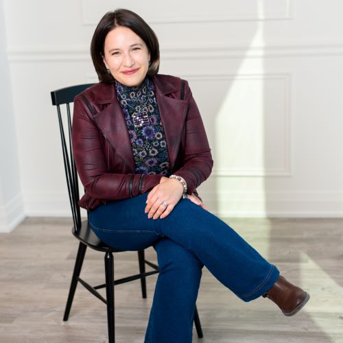 Sidura Ludwig, a light-skinned woman with short dark hair, sits in a chair with her legs crossed in front of a white wall. She smiles closed-mouth at the viewer.